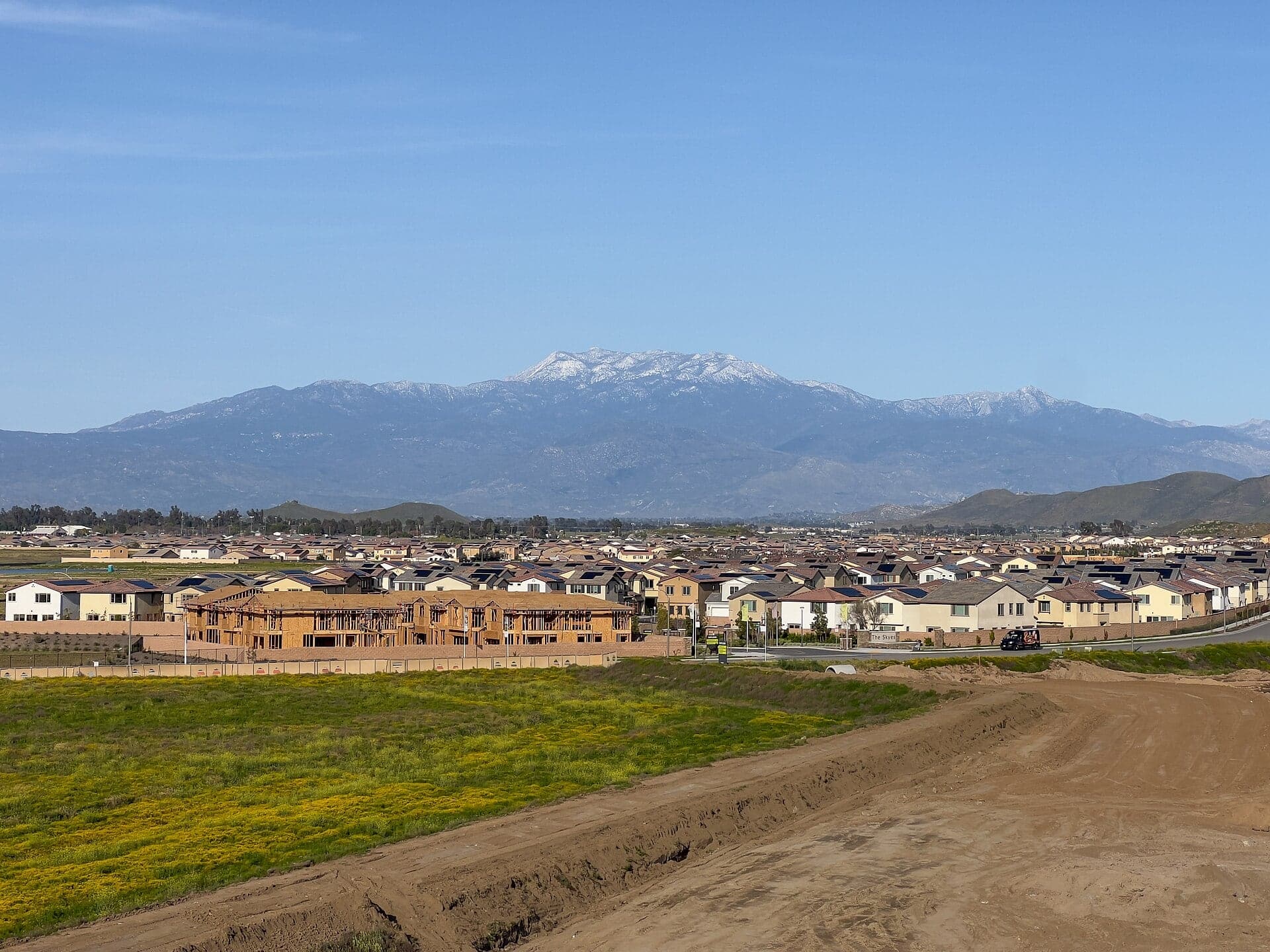 Wood-framed houses under construction at the edge of a new subdivision in Winchester, California, with rows of finished homes and the snow-capped San Jacinto Mountains behind